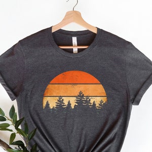 Peut inclure: Un t-shirt gris chiné foncé avec un graphique de coucher de soleil vintage représentant un soleil orange en demi-cercle avec trois bandes horizontales et une rangée de pins vert foncé devant.
