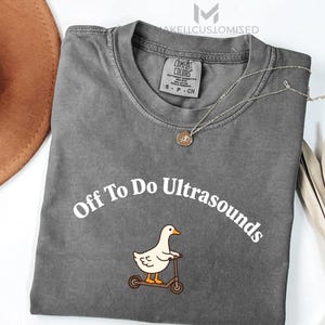 Peut inclure: Un t-shirt gris anthracite à manches courtes avec le texte blanc "Off To Do Ultrasounds" arqué au-dessus d'une illustration d'une oie blanche sur une trottinette marron. L'étiquette Comfort Colors au col indique la taille S-CH.