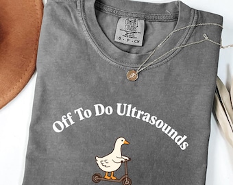 以下が含まれることがあります： チャコールグレーの半袖Tシャツ。白い文字で「Off To Do Ultrasounds」とアーチ状にプリントされ、その下に茶色のスクーターに乗った白いガチョウのグラフィックがある。首元のComfort ColorsのタグにはサイズS-CHと記載されている。