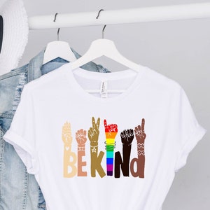 May include: White t-shirt featuring a graphic of hands in various skin tones forming the word "BE KIND". The hands are making the sign language symbol for "I love you".