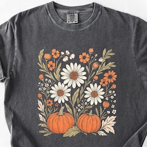 Peut inclure: T-shirt gris foncé avec un motif floral composé de marguerites blanches, de fleurs orange et de deux citrouilles orange. Le motif est centré, créant une esthétique automnale. Le t-shirt a des manches courtes et un col rond.