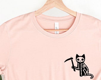 May include: A light pink t-shirt with a black skeleton cat holding a scythe printed on the front.