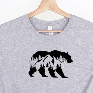 Puede incluir: Una camiseta gris con una silueta negra de un oso con montañas y árboles dentro del contorno del oso.