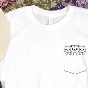 May include: White t-shirt with a black outline of three cats peeking out of a pocket on the front of the shirt.
