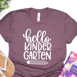 May include: A maroon t-shirt with the text "hello kindergarten" in white lettering and a white pencil graphic.