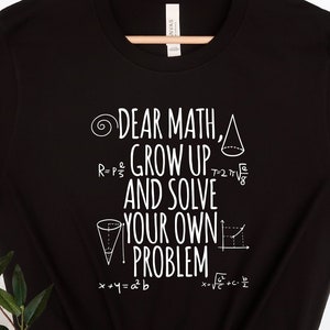 Peut inclure: T-shirt noir avec un texte blanc qui dit "Cher Math, grandis et résous tes propres problèmes" avec des équations mathématiques et des symboles entourant le texte.