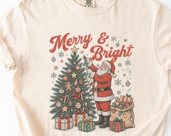 以下が含まれることがあります： クリスマスツリーを飾るサンタクロースのグラフィックがプリントされたクリーム色の半袖Tシャツ。イラストの上部には「Merry & Bright」という文字があり、包まれたギフトやオーナメントが入った袋が描かれています。