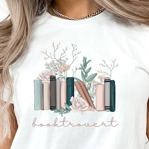May include: White t-shirt featuring a graphic of a stack of books with floral accents and the text "booktrovert".