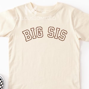 May include: A cream-coloured t-shirt with the text "BIG SIS" in brown block letters. The shirt is paired with black and white chequered slip-on shoes. Crayons and sunglasses are also in the image.