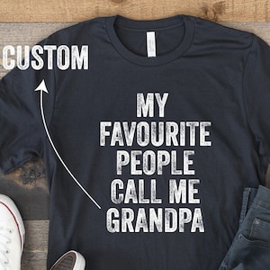 Puede incluir: Camiseta negra con texto blanco que dice "Mis personas favoritas me llaman Abuelo". La camiseta está puesta sobre una superficie de madera con un par de zapatillas blancas y unos vaqueros azules.