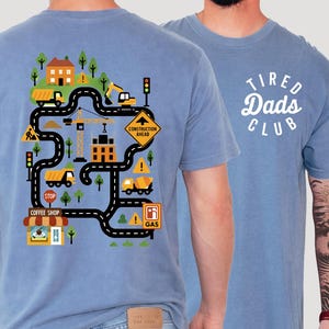 May include: Two light blue t-shirts. One has a colourful construction-themed graphic with a road, buildings, and vehicles. The other shirt features the text "Tired Dads Club" in a white, circular design. Both are casual wear.