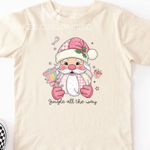 May include: A cream-coloured t-shirt with a Santa Claus graphic in pink and white, holding a lantern and a gift. The text "Jingle all the way" is below the graphic. Black and white checkered slip-on shoes are on the left.