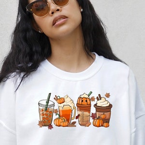 May include: White sweatshirt featuring a graphic of four autumn-themed drinks with pumpkins and leaves. The drinks include a pumpkin spice latte, a caramel apple cider, a pumpkin pie smoothie, and a pumpkin spice iced coffee.