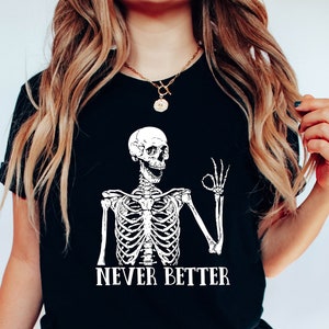K&ouml;nnte beinhalten: Ein schwarzes T-Shirt mit einem wei&szlig;en Skelett-Motiv, das einen Daumen hoch zeigt, und dem Text "NEVER BETTER".