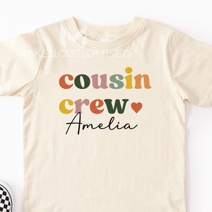 May include: A cream-coloured t-shirt featuring the words "cousin crew" in colourful letters and the name "Amelia" in a script font. A red heart is between "crew" and "Amelia". Black and white checkered slip-on shoes are on the left.