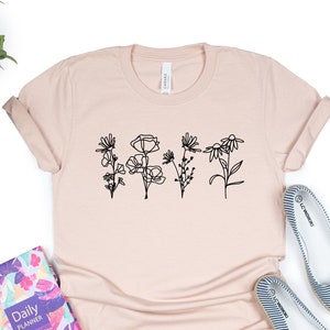May include: A light pink t-shirt with a black line drawing of five flowers on the front. The flowers are stylised and have a simple design.