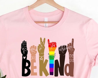 May include: A light pink t-shirt with the words "BE KIND" in colorful letters and hand gestures. The hands are in various skin tones and the rainbow-colored hand represents the LGBTQ+ community. The shirt is hanging on a wooden hanger.