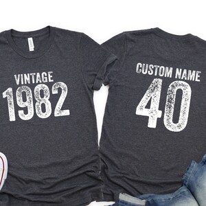 May include: Two dark gray t-shirts with white text. One shirt says "Vintage 1982" and the other says "Custom Name 40".