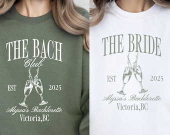 May include: Two green and white sweatshirts with a bachelorette party theme. The text on the green sweatshirt reads "The Bach Club Est 2025 Alyssa's Bachlorette Victoria, BC". The text on the white sweatshirt reads "The Bride Est 2025 Alyssa's Bachlorette Victoria, BC". Both sweatshirts feature a graphic of two champagne glasses clinking.