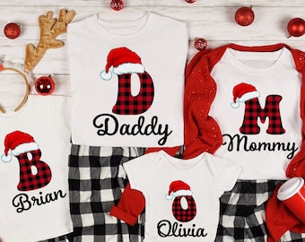 Könnte beinhalten: Ein passender Weihnachtspyjama für eine vierköpfige Familie mit rot-schwarzen Buffalo-Karopantalons und weißen Shirts mit einer roten Weihnachtsmütze und einem Buchstaben mit dem Namen "Daddy", "Mommy", "Brian" und "Olivia".
