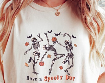 May include: A cream colored t-shirt with a graphic of three skeletons dancing and holding pumpkins and a trick-or-treat bag. The text "Have a Spooky Day" is printed below the skeletons.