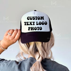 May include: A black and white trucker hat with a white mesh back. The hat has black text that reads "CUSTOM TEXT LOGO PHOTO" in a retro font.