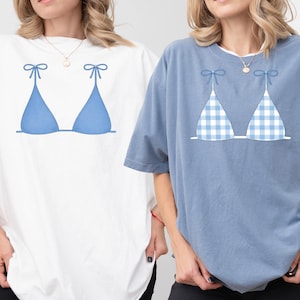 May include: Two oversized t-shirts, one white and one blue, each with a blue bikini top design. The white shirt has a solid blue bikini top, and the blue shirt has a blue and white gingham pattern.