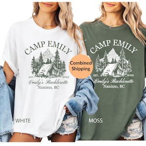 May include: Two t-shirts, one white and one moss green, featuring a camping design. The graphic includes the text "CAMP EMILY," "Emily's Bachelorette," and "Nanimo, BC." The white shirt is labelled "WHITE," and the green shirt is labelled "MOSS."
