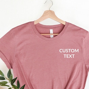 May include: A dusty rose coloured t-shirt with short sleeves and a crew neck. The shirt has the text "CUSTOM TEXT" printed on the front in white.