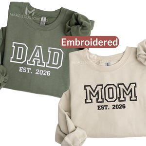 Peut inclure: Deux sweatshirts brodés, un vert olive et un beige. Le sweat-shirt vert porte l'inscription "DAD EST. 2026" en lettres blanches. Le sweat-shirt beige porte l'inscription "MOM EST. 2026" en lettres noires. Les deux ont des manches longues.
