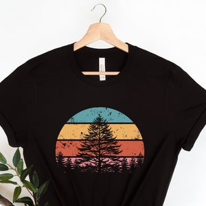 Peut inclure: T-shirt noir avec un graphique de style vintage d'un pin devant un coucher de soleil. Le coucher de soleil est un demi-cercle avec des rayures bleues, turquoise, orange, rouge et rose.