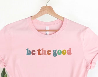 Könnte beinhalten: Rosa T-Shirt mit dem Text "be the good" in bunten Retro-Lettern.