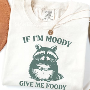 May include: Cream-coloured t-shirt featuring a raccoon graphic and the text "IF I'M MOODY GIVE ME FOODY" in green. A gold necklace and a brown hat are also visible.