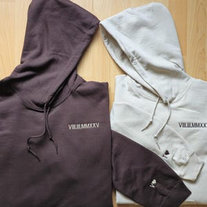 May include: Two hooded sweatshirts, one in a deep brown and the other in a light beige. Both have the Roman numeral "VIII.III.MMXXV" embroidered on the chest. The brown sweatshirt has a matching sleeve with a small embroidered heart.