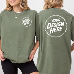 May include: An olive green t-shirt featuring the text "YOUR DESIGN HERE" in a white circle on the front and back. The shirt has short sleeves and a relaxed fit. The model is wearing black shorts.