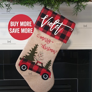 May include: A festive Christmas stocking featuring a red and black check cuff. The tan stocking has a red truck design carrying a Christmas tree. The name "Violet" is written above "merry christmas".