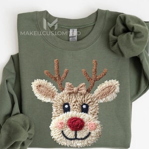 May include: An olive green sweatshirt with a textured reindeer face. The reindeer has brown antlers, a red nose, and a small bow. The text "MAKELLCUSTOMIZED" is visible. A festive, cosy design.