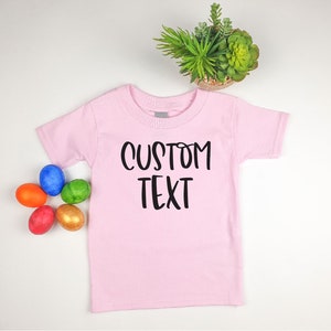 Puede incluir: Camiseta rosa para beb&eacute; con texto negro que dice "CUSTOM TEXT". La camiseta est&aacute; puesta sobre una superficie blanca con cinco huevos de Pascua de colores.
