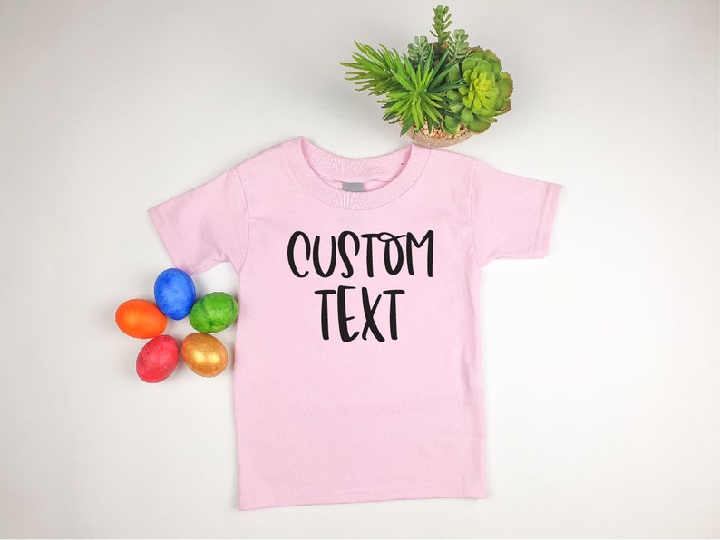 Puede incluir: Camiseta rosa para beb&eacute; con texto negro que dice "CUSTOM TEXT". La camiseta est&aacute; puesta sobre una superficie blanca con cinco huevos de Pascua de colores.