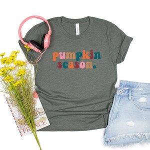 Może przedstawiać: Zielony t-shirt z napisem "Pumpkin season." w kolorowych literach. Koszulka leży płasko na białym tle z r&oacute;żowymi słuchawkami, bukietem ż&oacute;łtych kwiat&oacute;w i książką.