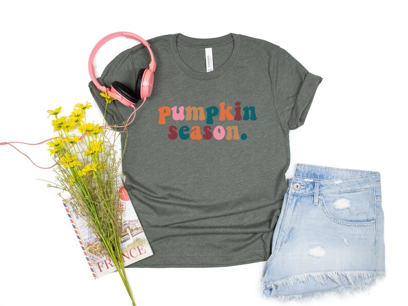 Może przedstawiać: Zielony t-shirt z napisem "Pumpkin season." w kolorowych literach. Koszulka leży płasko na białym tle z r&oacute;żowymi słuchawkami, bukietem ż&oacute;łtych kwiat&oacute;w i książką.