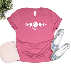 May include: A pink t-shirt with a white graphic of a moon phase design. The design features a diamond shape with a full moon in the center and crescent moons on either side.
