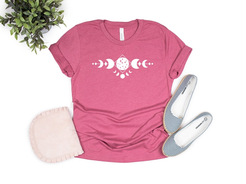 May include: A pink t-shirt with a white graphic of a moon phase design. The design features a diamond shape with a full moon in the center and crescent moons on either side.