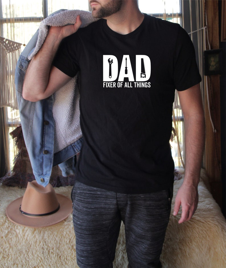 May include: Black t-shirt with white text that reads "DAD FIXER OF ALL THINGS". The text is surrounded by a wrench, screwdriver, and saw.