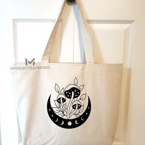 May include: A white canvas tote bag with a black design of mushrooms growing under a crescent moon with stars. The moon has three phases.