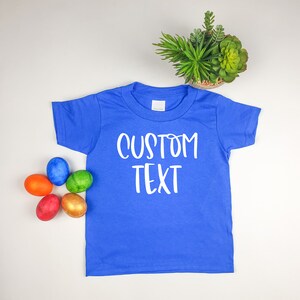 Puede incluir: Una camiseta azul real de manga corta con el texto "CUSTOM TEXT" impreso en blanco en la parte delantera. La camiseta est&aacute; colocada sobre una superficie blanca con cinco huevos de Pascua coloridos en la esquina inferior izquierda.