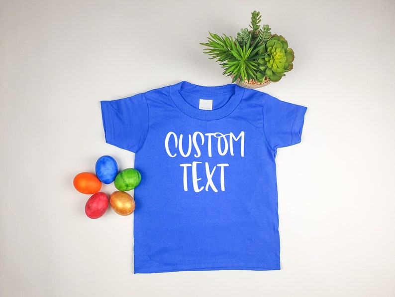 Puede incluir: Una camiseta azul real de manga corta con el texto "CUSTOM TEXT" impreso en blanco en la parte delantera. La camiseta est&aacute; colocada sobre una superficie blanca con cinco huevos de Pascua coloridos en la esquina inferior izquierda.