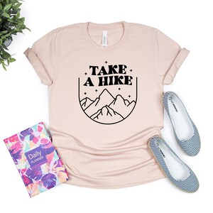 May include: A light pink t-shirt with a black graphic design that says "TAKE A HIKE" with a mountain range and stars inside a circle.
