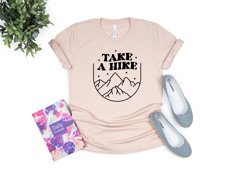 May include: A light pink t-shirt with a black graphic design that says "TAKE A HIKE" with a mountain range and stars inside a circle.