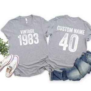 K&ouml;nnte beinhalten: Zwei graue T-Shirts mit wei&szlig;em Text. Das vordere Shirt sagt "Vintage 1983" und das hintere Shirt sagt "Custom Name 40".